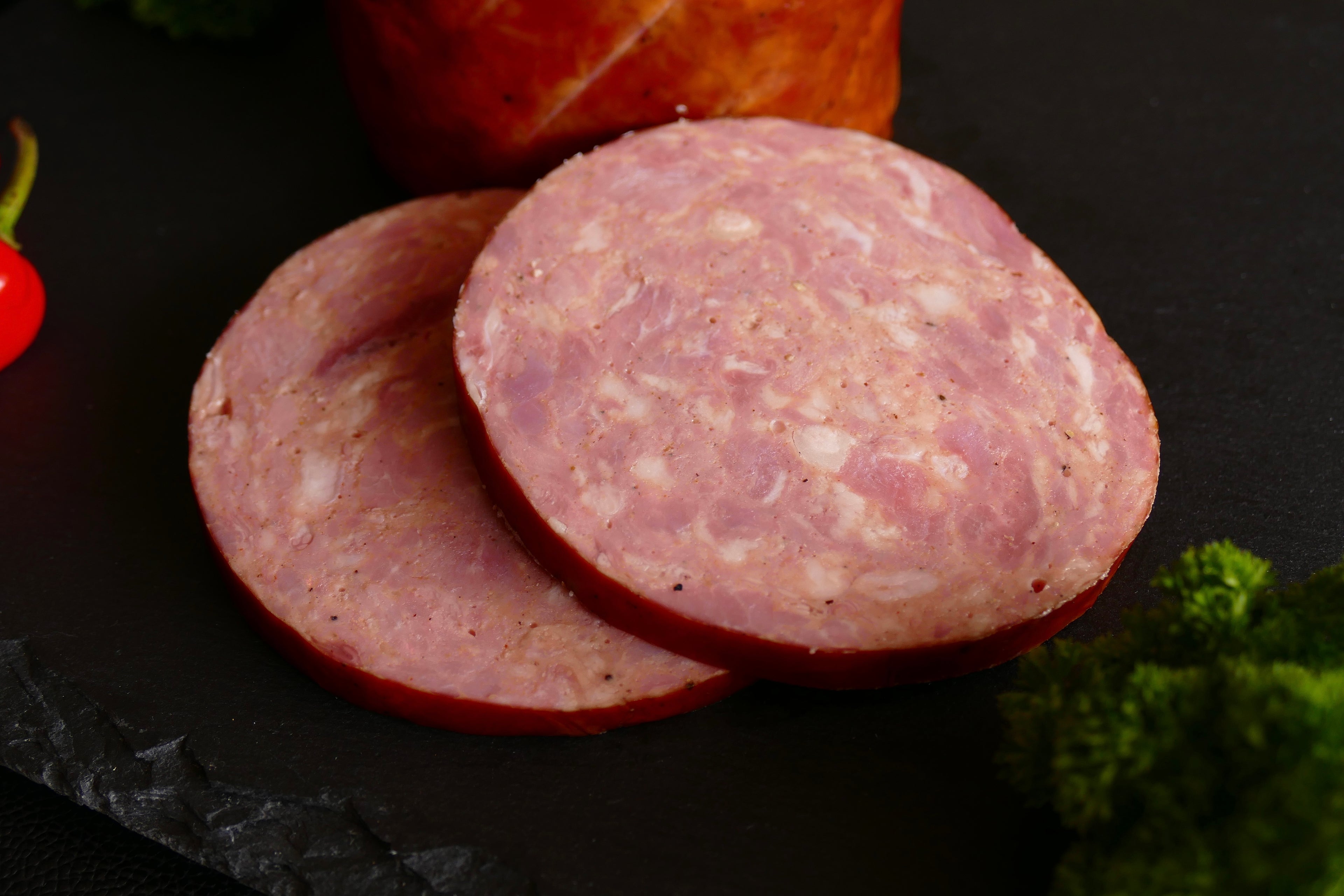 Two thick slices of pink cured sausage or salami with visible fat marbling, arranged on a dark surface with a whole sausage roll blurred in the background and small garnish accents at the edges.